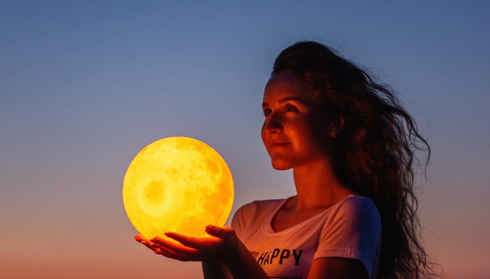 A young woman holds the full moon in her hands against the backdrop of a red sunset, understanding the full moon effects and impacts the full moon has on humans