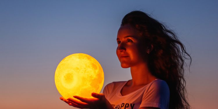 A young woman holds the full moon in her hands against the backdrop of a red sunset, understanding the full moon effects and impacts the full moon has on humans