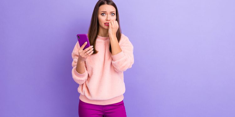 Photo of pretty blogger lady with phone axniety hold telephone not believe unfollowers, number biting fingers fear eyes wear casual fur pullover trousers isolated purple color background