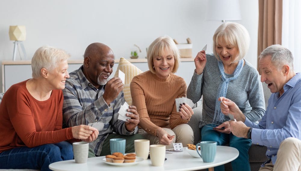 Multiethnic group of happy senior friends men and women in casual sitting on couch, having conversation and playing cards while drinking tea with cookies, chilling together at home after choosing assisted living