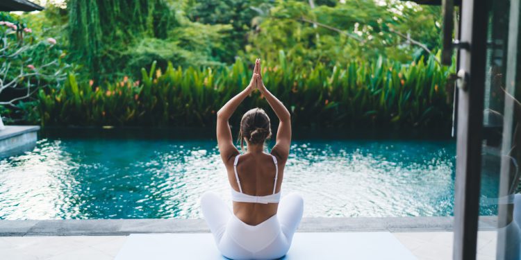 Back view of peaceful female in sportswear sitting on mat near pool and exotic garden praying during namaste zen practice at wellness retreats