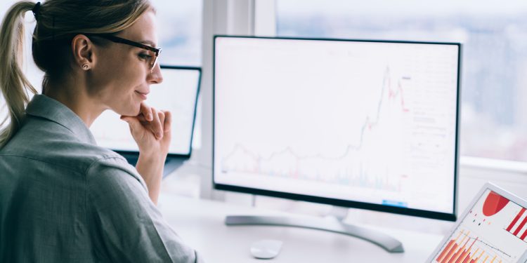 Side view of female financial analyst touching chin while pointing with stylus at tablet screen with chart near desktop computer at work, needing mindfulness tips for traders