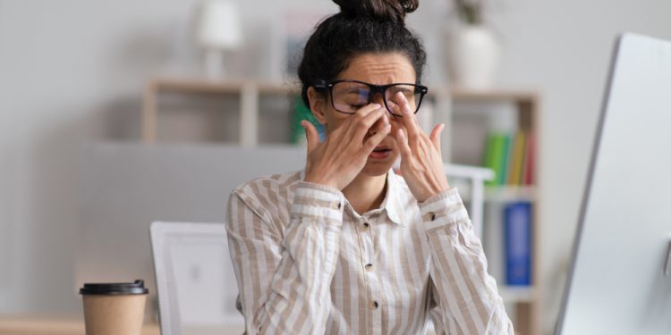 Female office worker in glasses rubbing tired eyes, exhausted from overworking, sitting at workplace in office, copy space. Young businesswoman feeling eye fatigue or pain, needing to learn how to deal with stress as a 911 dispatcher